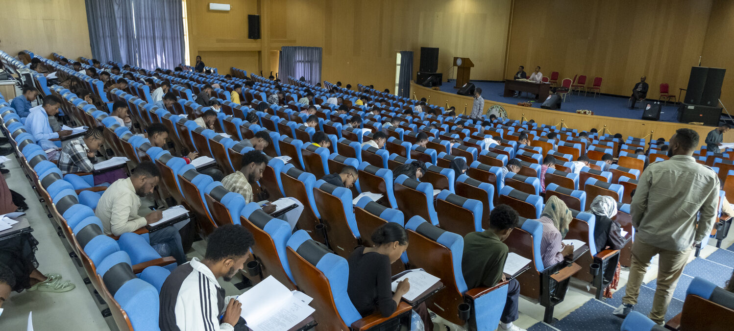 Studierende sitzen in Reihen von blauen Stühlen in einem großen Hörsaal und schreiben eine Prüfung.