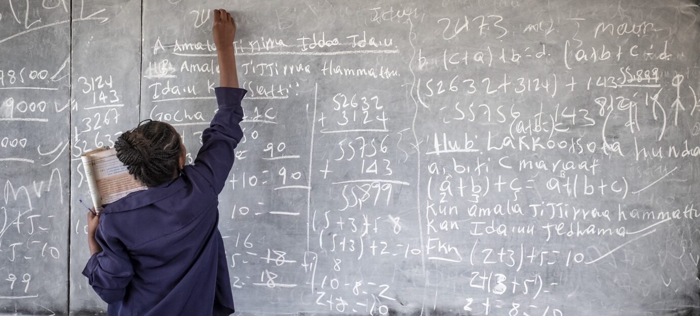 Ein Schüler schreibt mathematische Gleichungen auf eine Kreidetafel und hält dabei ein Notizbuch in der Hand.