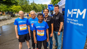Fünf Männer in blauen T-Shirts von Menschen für Menschen lächeln sich bei einer Veranstaltung im Freien an.