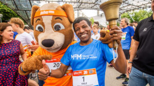 Ein Mann im blauen Hemd mit einer Glocke steht bei einer Laufveranstaltung neben einem Hundemaskottchen.