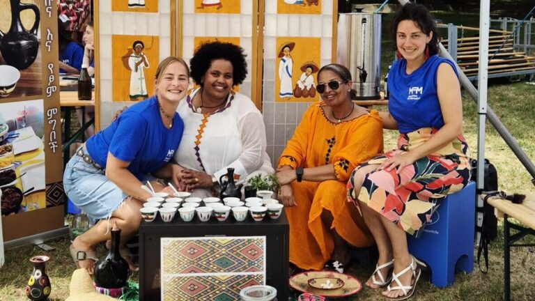 Vier Frauen lächeln an einem Kaffeestand; zwei tragen blaue Hemden, zwei tragen Kleider, bei einer Veranstaltung.