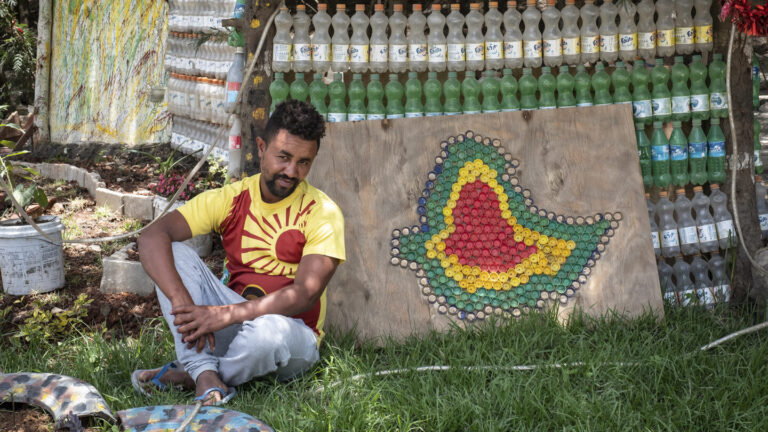 Mann im Gras neben einer Karte in Form eines afrikanischen Flaschenverschlusses, im Hintergrund aufgereihte Plastikflaschen.