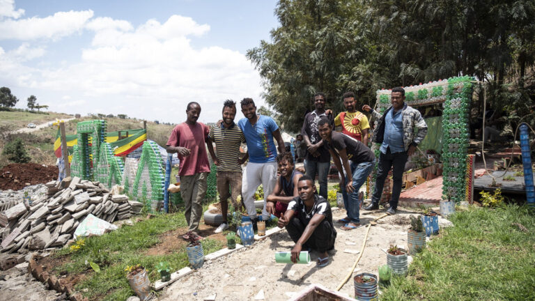 Eine Gruppe von Menschen lächelt im Freien in der Nähe von bunten Strukturen und gestapelten Lieferungen.