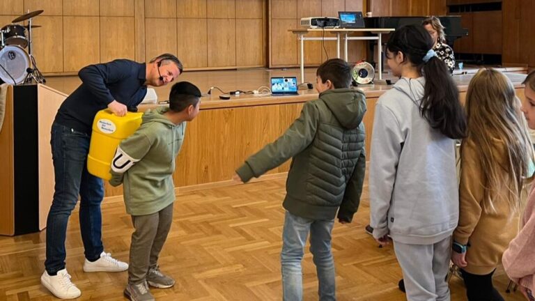 Ein Mann hält spielerisch einen gelben Eimer an einen lächelnden Jungen, andere Kinder in der Nähe schauen zu und lächeln.