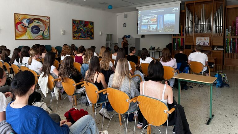 Schüler in einem Klassenzimmer sehen sich eine Präsentation an, die auf eine Leinwand im vorderen Bereich projiziert wird.