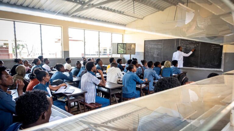 Ein Lehrer schreibt auf einer Kreidetafel, während Schüler in blauen Uniformen an den Tischen in einem Klassenzimmer sitzen.