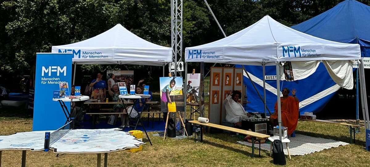 Zwei weiße Zelte von Mensen für Menschen bei einer Freiluftveranstaltung mit Auslagen und Besuchern.