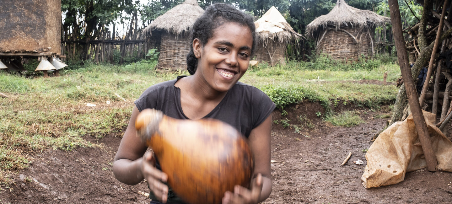 Junge Frau hält lächelnd einen großen Flaschenkürbis im Freien, im Hintergrund sind runde Hütten zu sehen.
