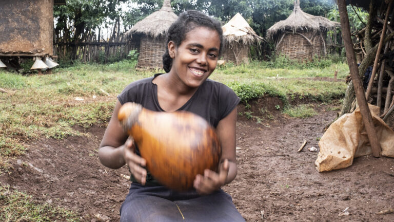 Eine Frau hält lächelnd einen großen Flaschenkürbis im Freien; im Hintergrund sind traditionelle Hütten zu sehen.