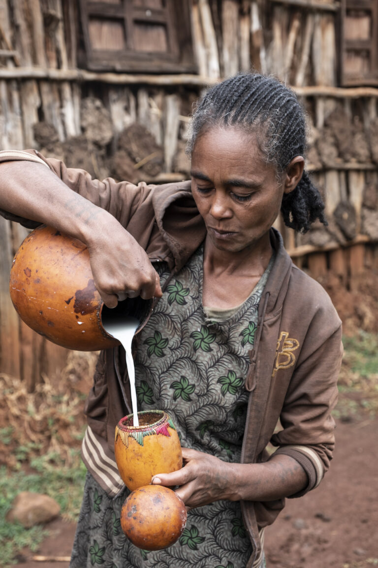 Eine Frau gießt Milch zwischen Kürbissen vor einer rustikalen Mauer ein.