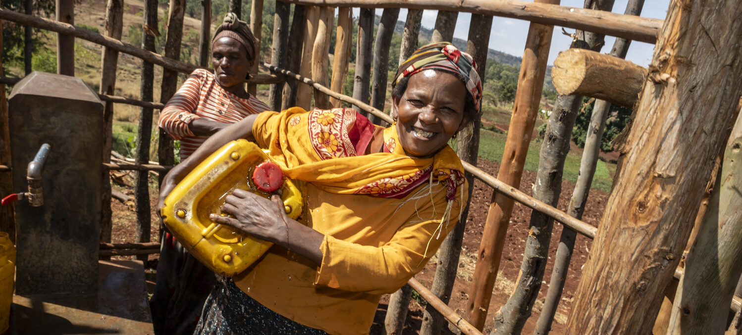 Zwei Frauen lächeln, während sie an einem Brunnen im Freien gelbe Wasserbehälter füllen.