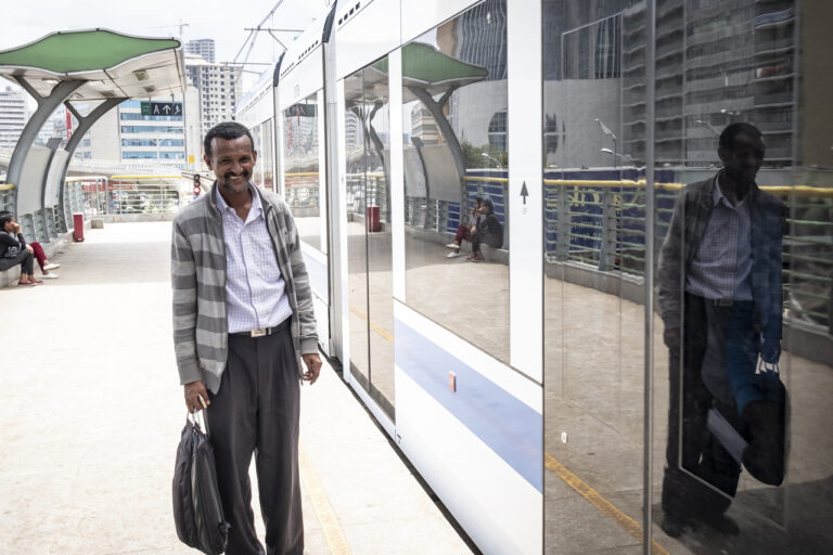 Ein lächelnder Mann mit einer Tüte in der Hand steht an einem Zug in einem modernen Bahnhof im Freien.