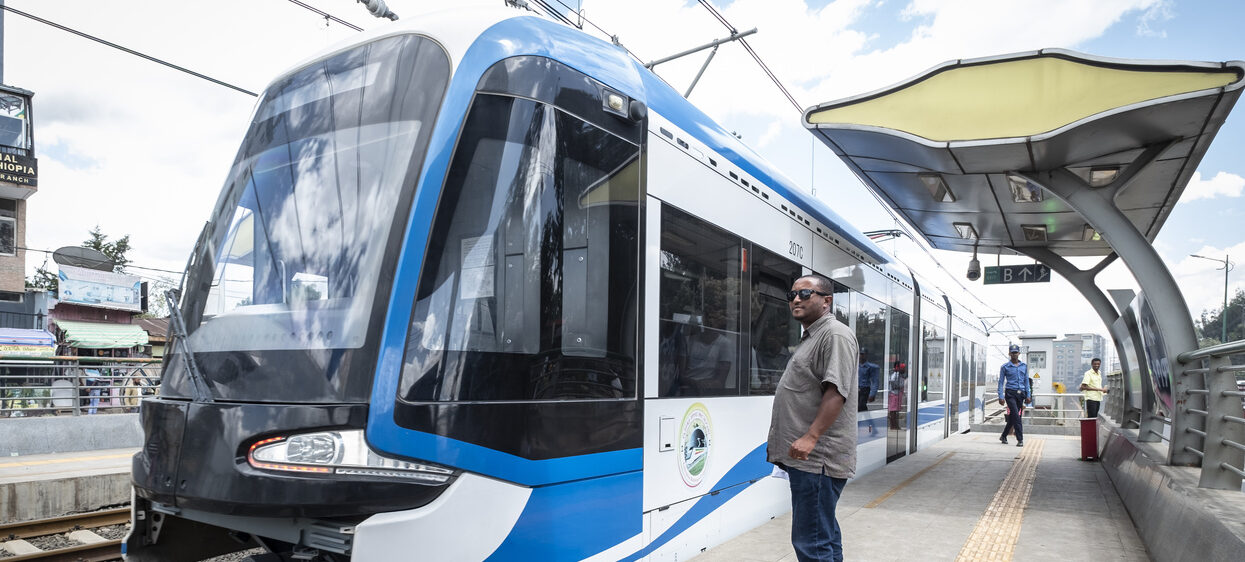 Blauer und weißer Stadtbahnzug an einer Außenstation mit wartenden Menschen in der Nähe.