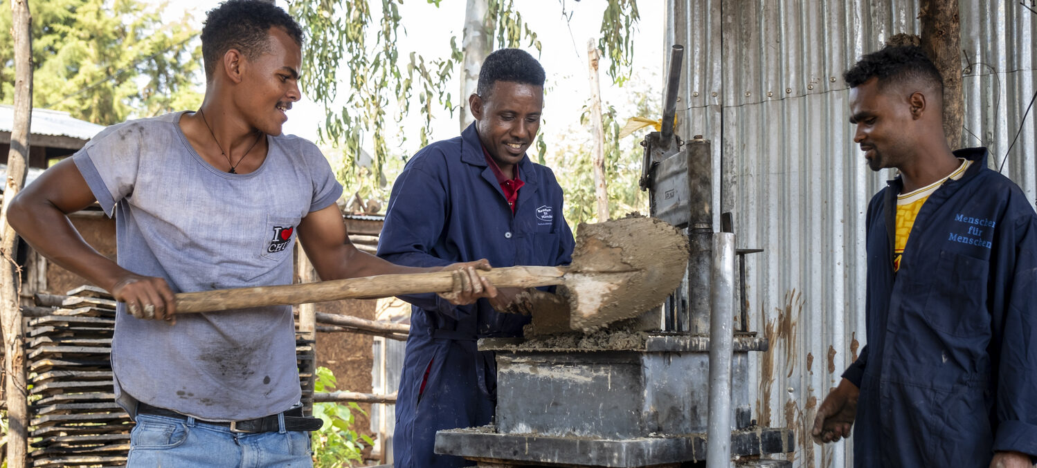 Drei Männer mischen unter freiem Himmel mit einer Schaufel und einer Maschine nassen Zement und arbeiten dabei zusammen.