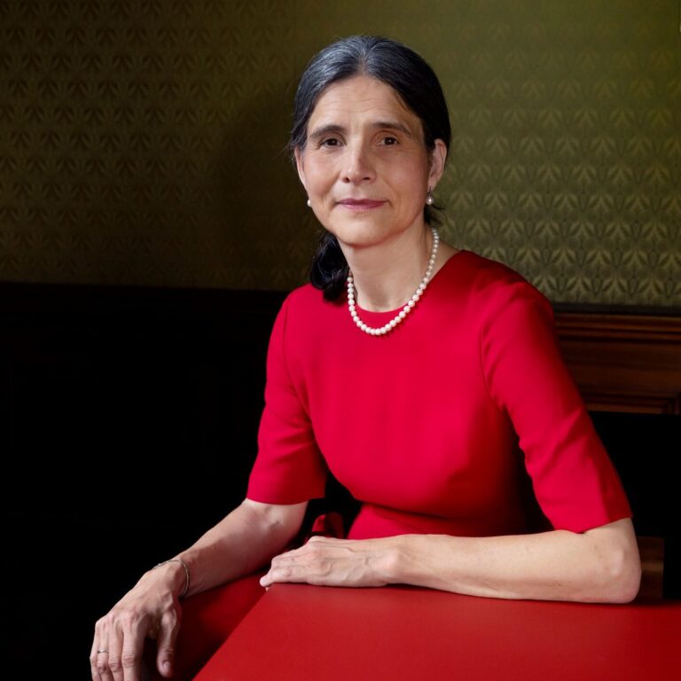 Frau in rotem Kleid mit Perlenkette sitzt an einem roten Tisch und blickt in die Kamera.