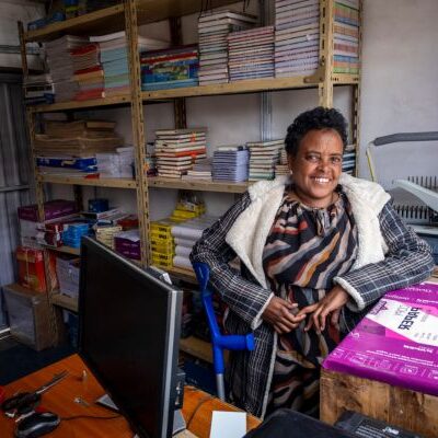 Lächelnde Frau in einem Büro, hinter ihr Regale mit Büchern und Material.