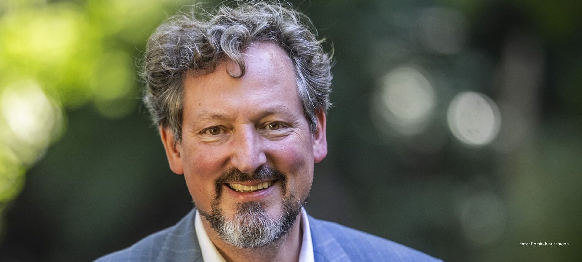Ein lächelnder Mann mit lockigem Haar und Bart im Anzug, im Freien, steht für eine glückliche Spenderunterstützung.