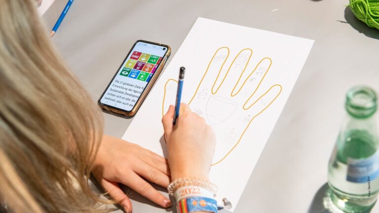 Person skizziert Hand auf Papier; Telefon mit Spenden-App-Symbolen und Wasserflasche liegen auf dem Tisch.