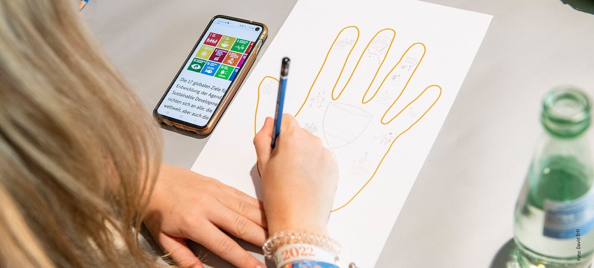 Jemand skizziert eine Hand auf Papier an einem Tisch, neben einem Telefon und einer Glasflasche.