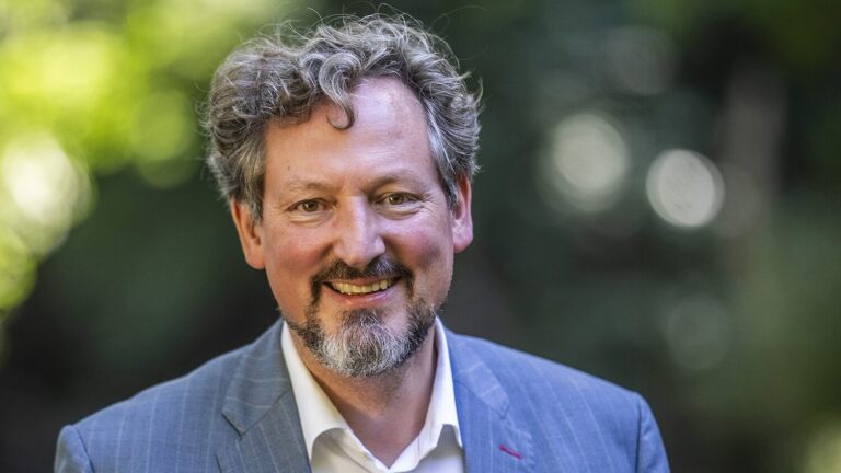 Lächelnder Mann mit lockigem Haar und Bart in einem blauen Anzug, der draußen auf der NGO-Webshop-Seite steht.