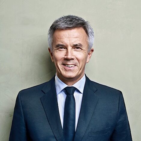 Ein freundlicher Mann in dunklem Anzug und Krawatte steht vor einem schlichten, hellen Hintergrund und lächelt freundlich.