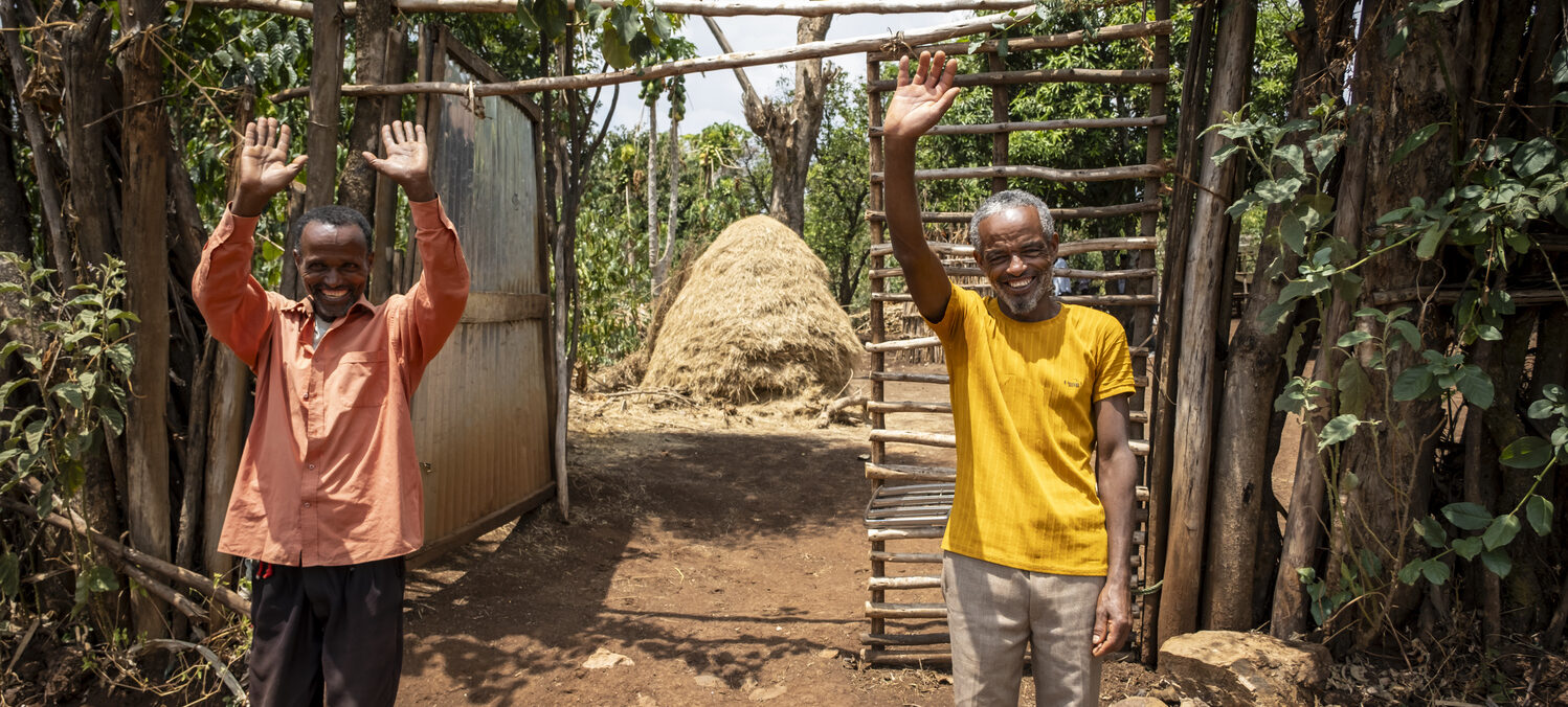 Ein lächelnder Mann winkt vor einem Holzgebäude und bittet um Unterstützung für den NGO-Spenden-Webshop.
