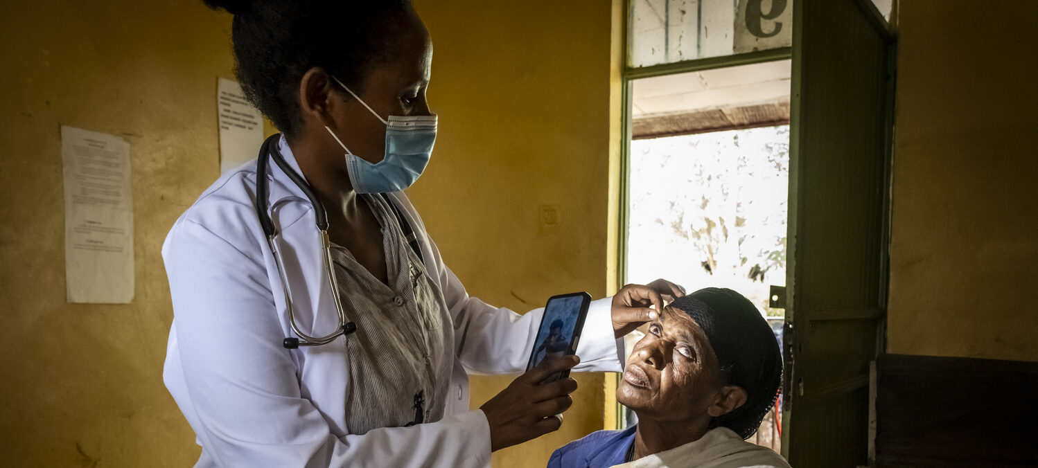 Maskierter Arzt überprüft das Auge eines Patienten mit einem Smartphone in einer Klinik; Bild für NGO-Spendenwebshop.