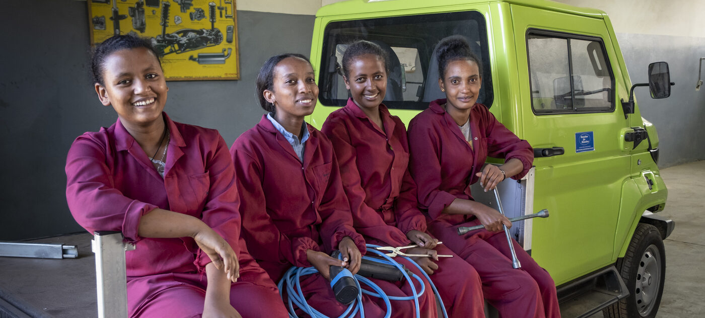 Vier lächelnde Frauen in roten Overalls sitzen mit Werkzeugen neben einem grünen Fahrzeug in einer Werkstatt.
