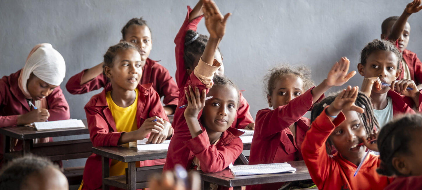 Kinder in roten Uniformen heben die Hände an den Tischen in einem Klassenzimmer, bereit zu lernen und mitzumachen.