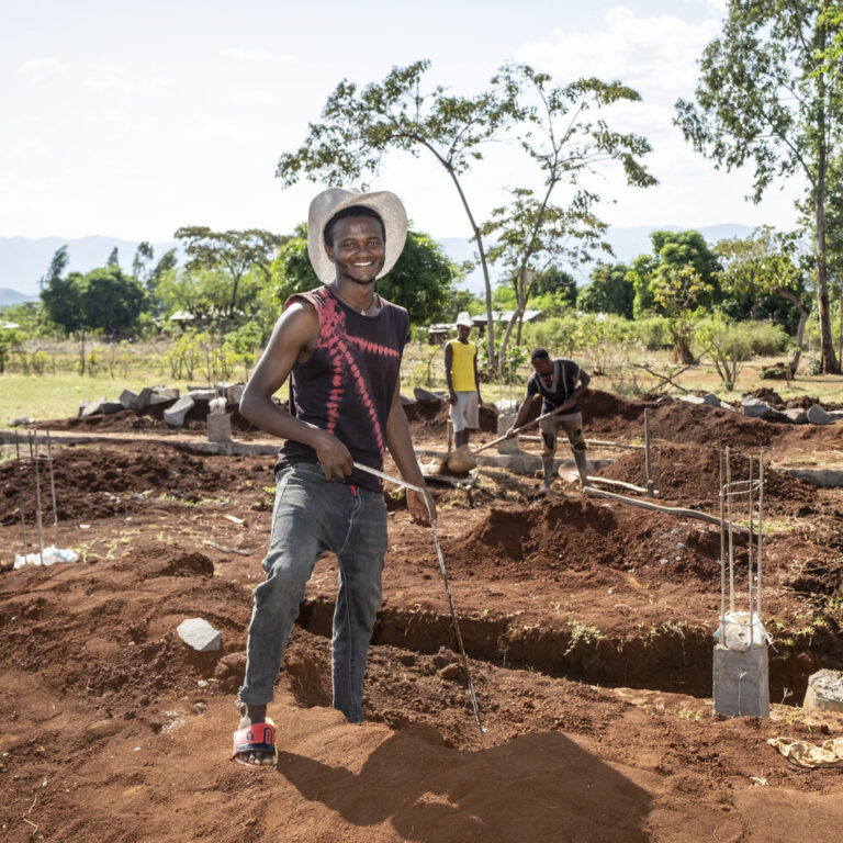 Lächelnder Mann mit Hut auf einer Baustelle, mit Menschen, die im Hintergrund arbeiten.