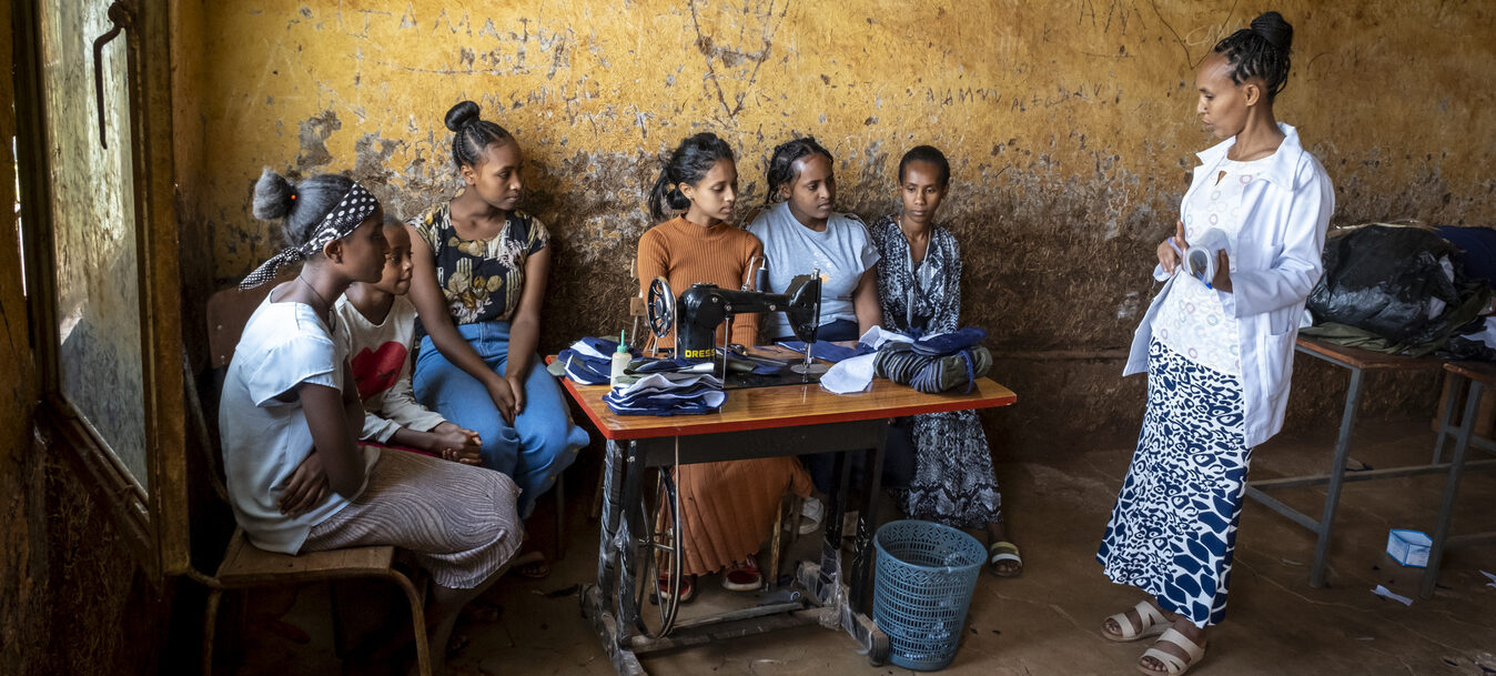 Sieben Frauen versammeln sich in einem rustikalen Raum, sechs sitzen an einer Nähmaschine, eine steht und spricht.