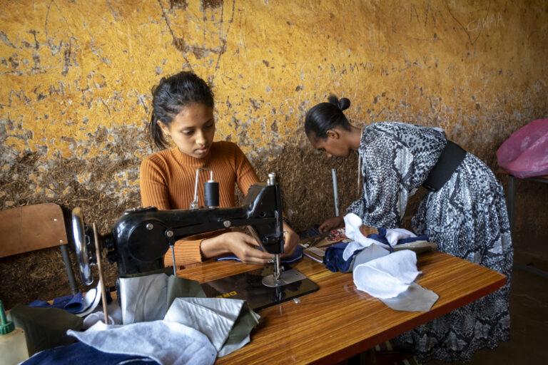Zwei Frauen in einem rustikalen Raum an einer Nähmaschine am Stoff zusammennähen.