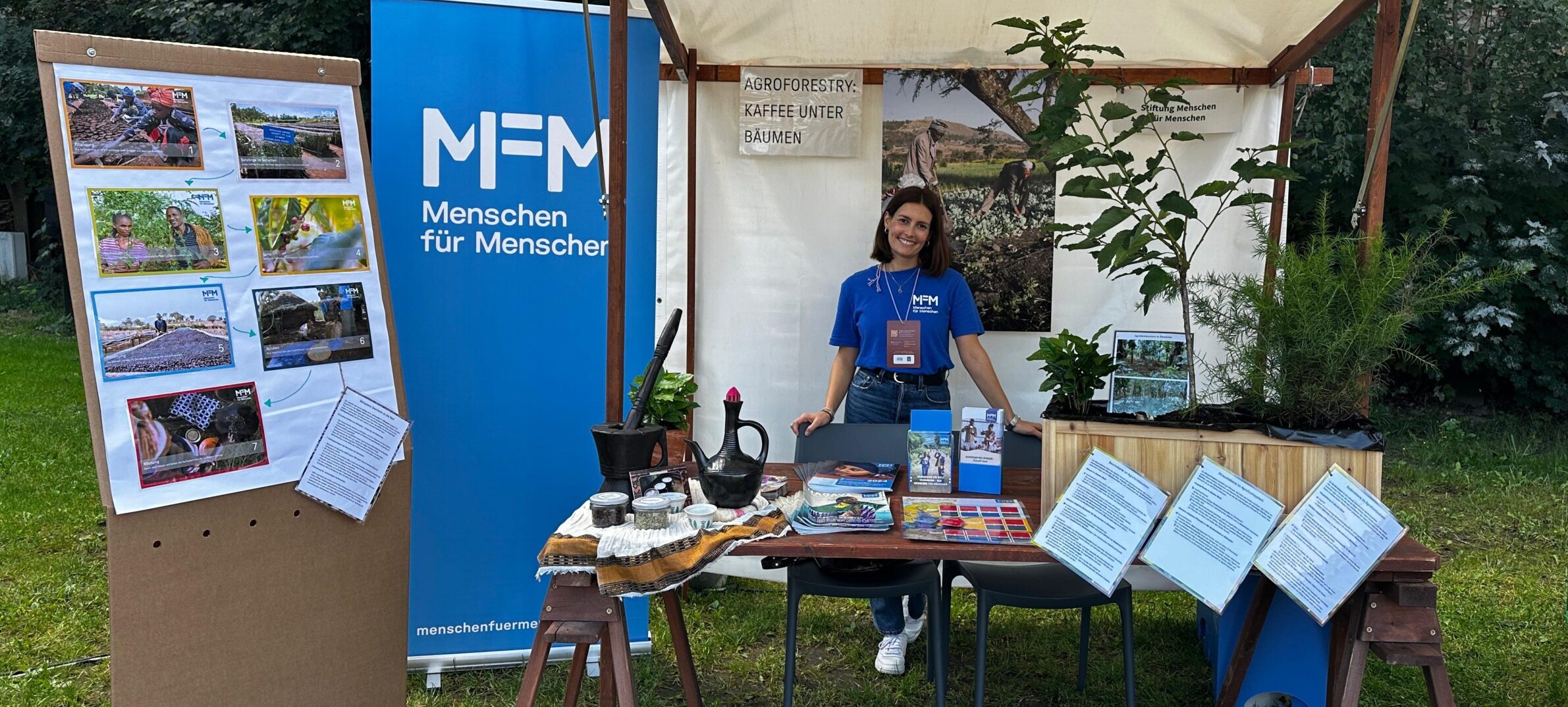 Frau am Stand mit Pflanzen, Fotos und Bannern von Menschen für Menschen bei einer Open-Air-Spendenaktion.