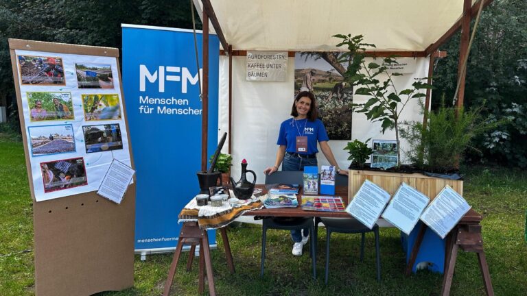 Eine Frau an einem Infostand von Menschen für Menschen mit Broschüren, die zu Spenden für den NGO-Webshop aufrufen.