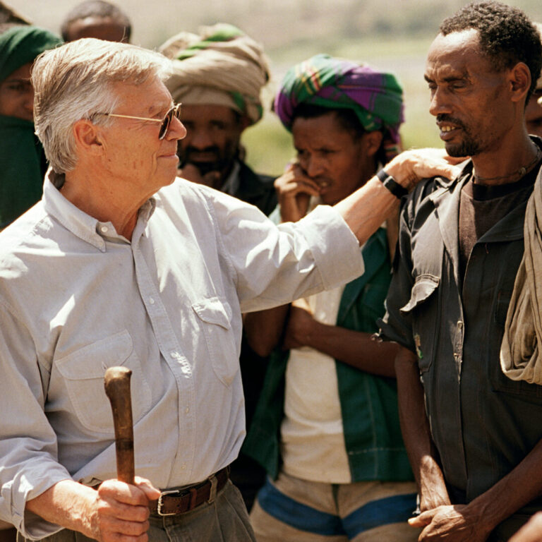 Ein älterer Mann mit Sonnenbrille stützt einen anderen im Freien, der einen Holzstock inmitten einer kleinen Gruppe hält.
