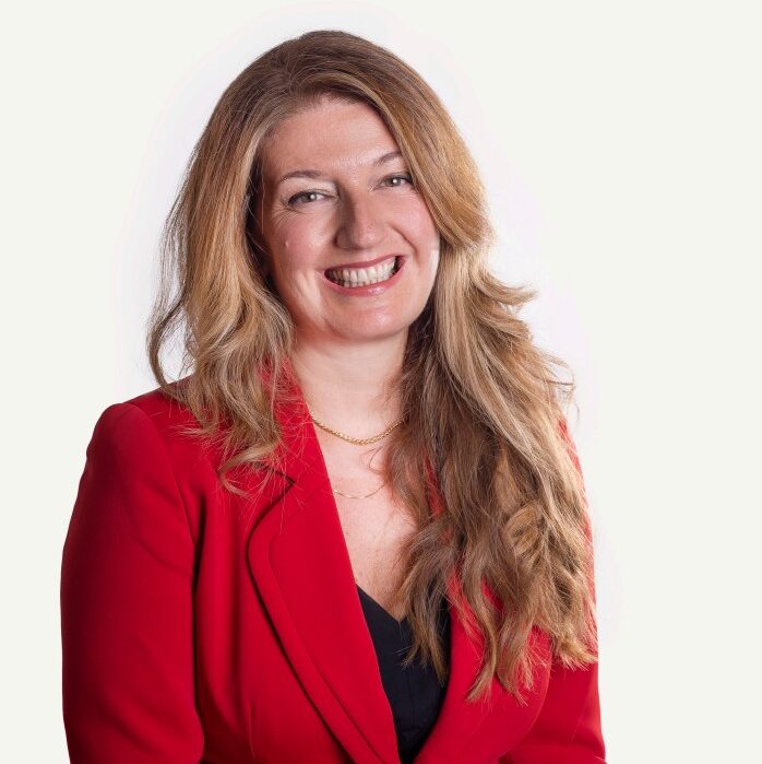 Lächelnde Frau mit langen gewellten Haaren in einem roten Blazer, die vor einem schlichten weißen Hintergrund posiert.