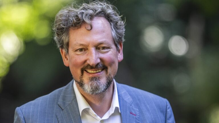 Lächelnder Mann mit lockigem Haar und Bart in einem blauen Anzug, der draußen steht und hinter sich Grünpflanzen hat.