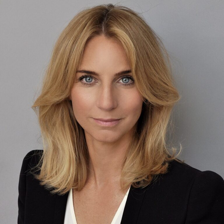 Blonde Frau mit schulterlangen Haaren in einem schwarzen Blazer, posiert vor einem schlichten grauen Hintergrund.