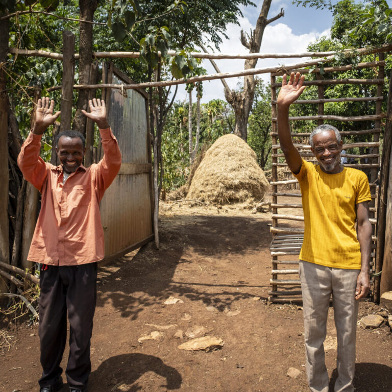Zwei Männer vor einem Tor lächeln und winken mit erhobenen Händen, wirken fröhlich und einladend.