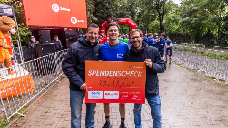 Drei lächelnde Männer mit einem Scheck über 60.000 bei einer Wohltätigkeitsveranstaltung im Freien.