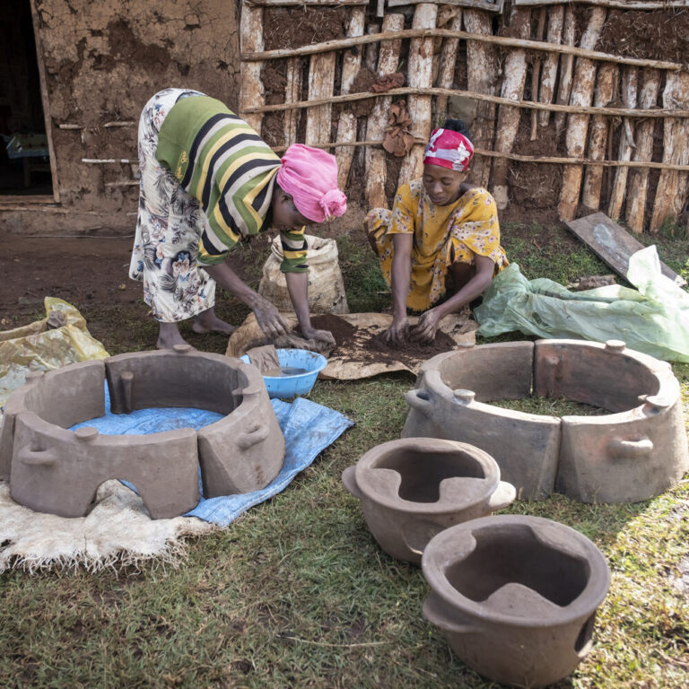 Zwei Frauen knien im Freien vor einem rustikalen Gebäude mit Lehmwänden und formen Lehmtöpfe, um Öfen für Spenden herzustellen.