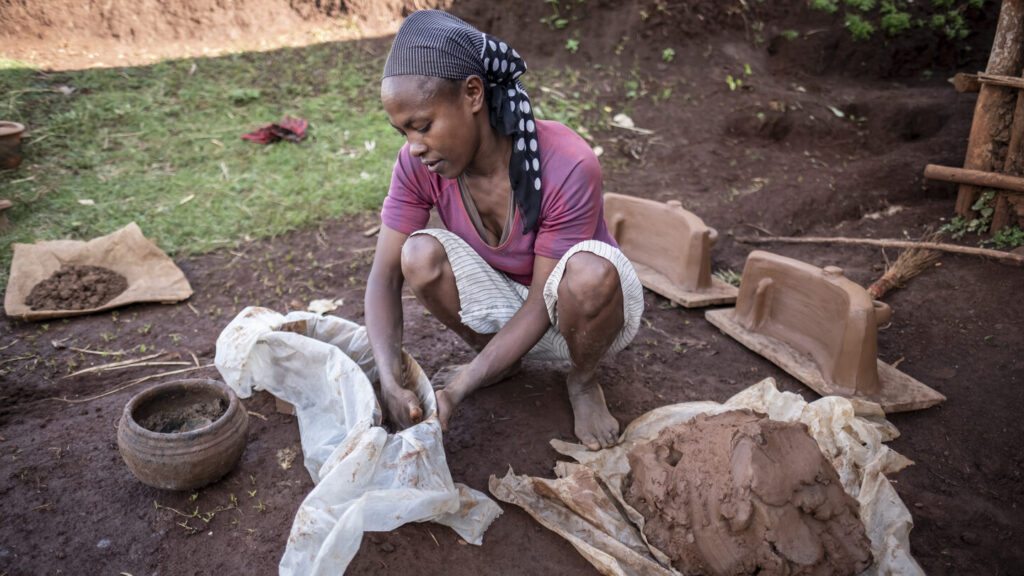 Eine Frau kniet im Freien und formt mit der Hand Ton; neben ihr liegen Töpferwaren und Handwerkszeug.