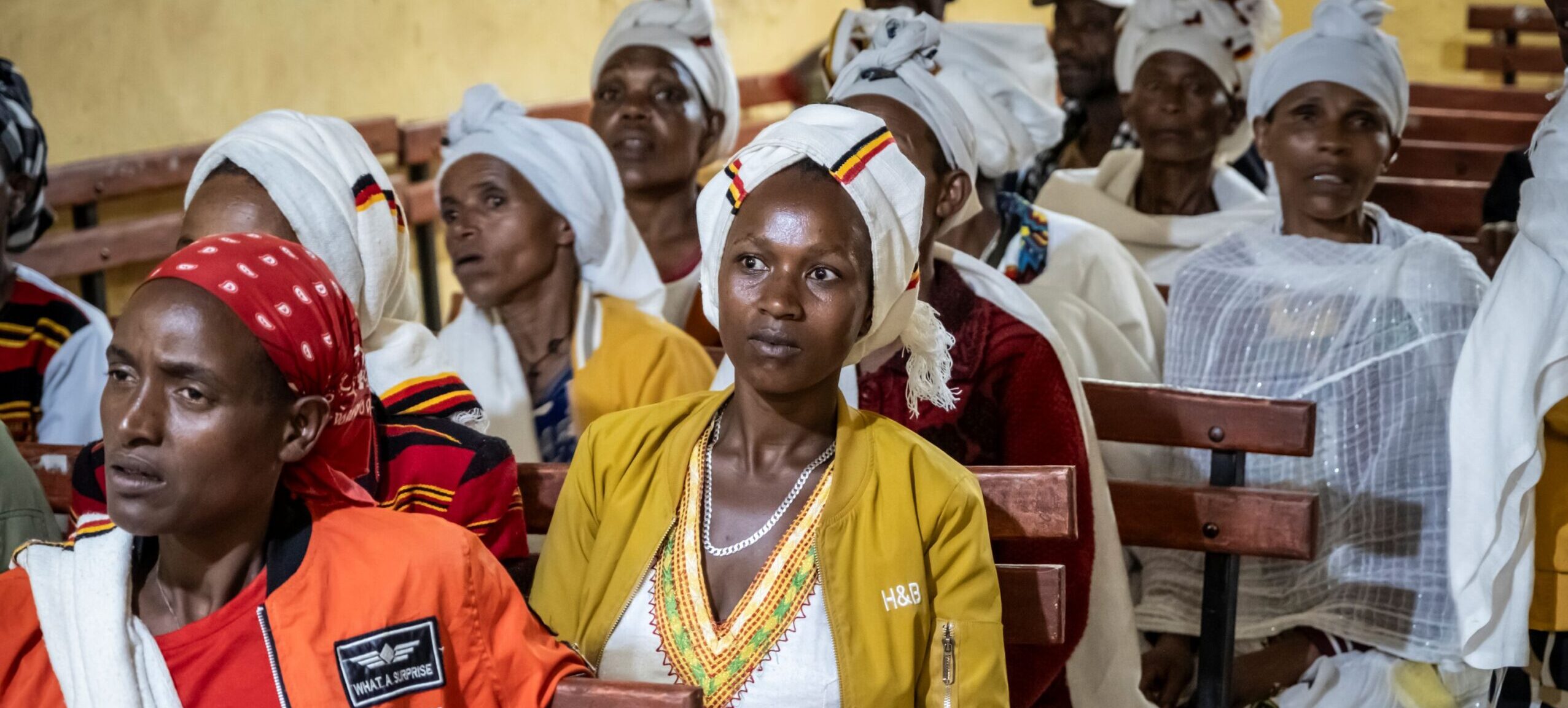 Frauen in farbenfrohen Kleidern und mit Kopftüchern sitzen auf Bänken, mit dem Gesicht nach vorne, in einer Gemeinschaft.