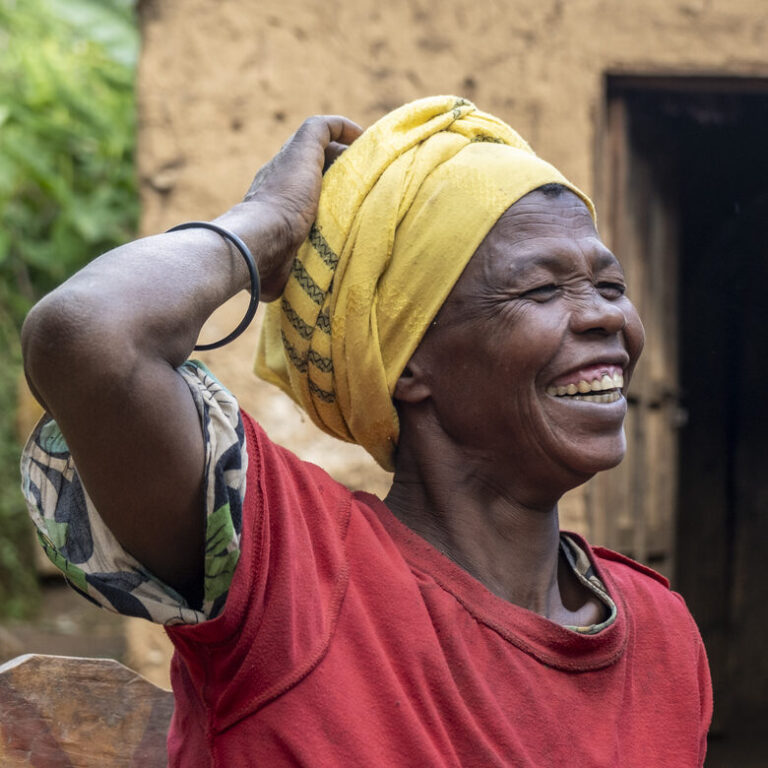 Frau mit gelbem Kopftuch und rotem Hemd lächelt im Freien, die Hand auf den Kopf gestützt.