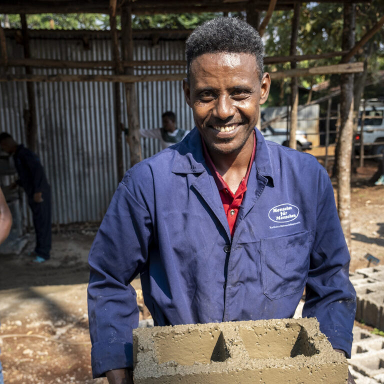 Arbeiter in blauem Overall lächelt und hält einen Betonblock auf einer Baustelle. Spenden Sie zur Unterstützung.