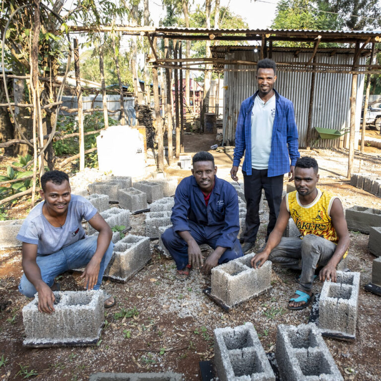 Vier Männer bauen mit Betonblöcken im Freien in der Nähe von Schuppen und Bäumen, um ein Gemeinschaftsprojekt zu unterstützen.