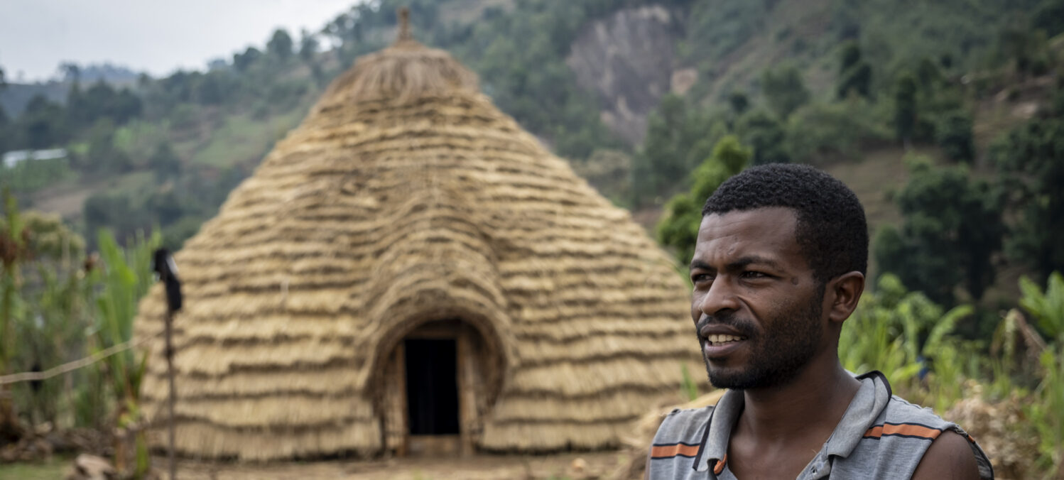 Ein Mann steht vor einer strohgedeckten Hütte, hinter der er Berge und Bäume sieht.