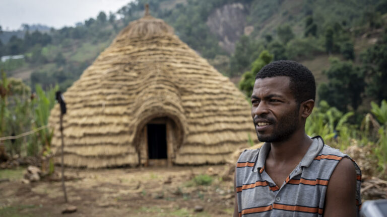 Ein Mann steht an einer runden, strohgedeckten Hütte vor einer grünen Hügellandschaft, die eine ländliche Gemeinschaft unterstützt.