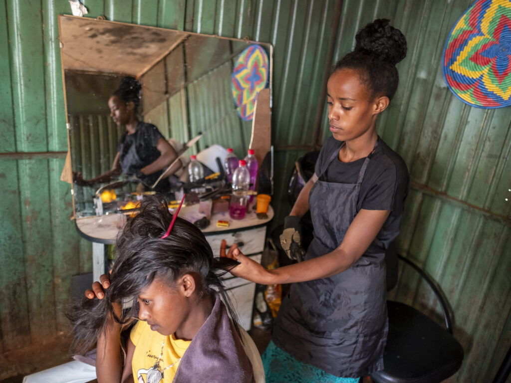 Zwei Frauen in einem kleinen Salon mit einer grünen Metallwand; die eine frisiert die Haare der anderen.
