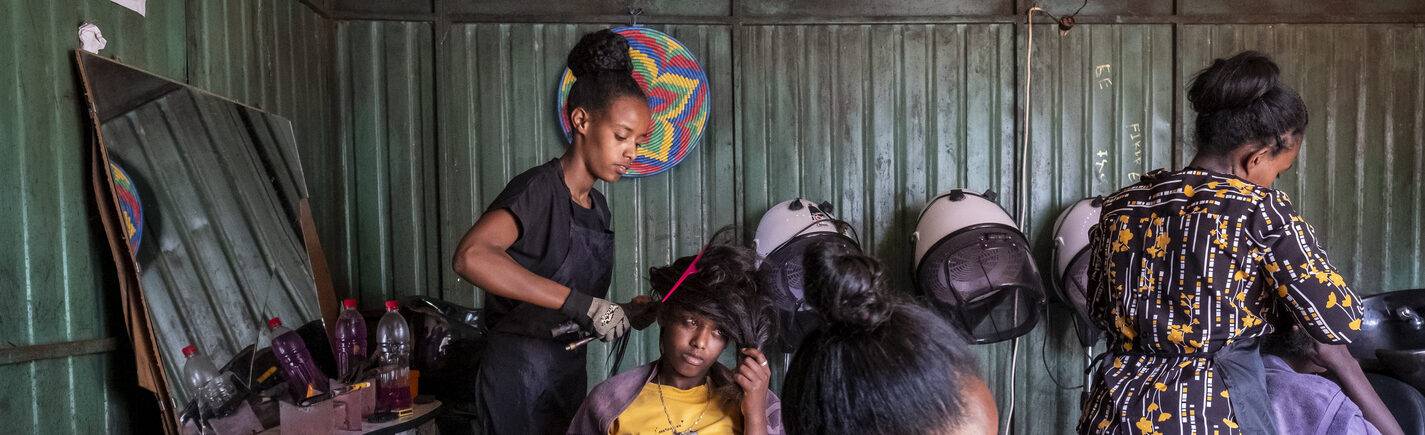 Drei Frauen in einem Friseursalon; eine stylt einer anderen die Haare, während eine dritte Frau im Hintergrund arbeitet.